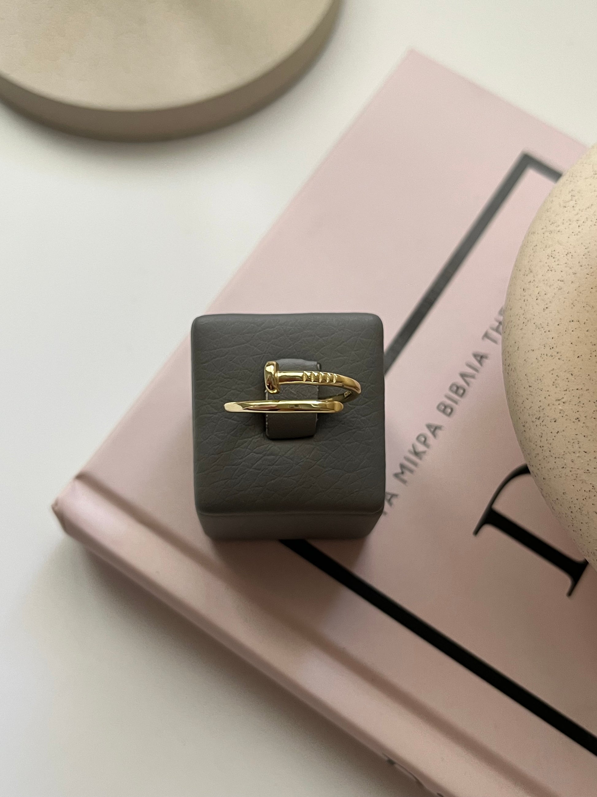 Χρυσό δαχτυλίδι σε σχέδιο καρφιού (nail ring) της Alista Jewels τοποθετημένο σε γκρι βάση πάνω σε βιβλίο – Μοντέρνο γυναικείο κόσμημα.